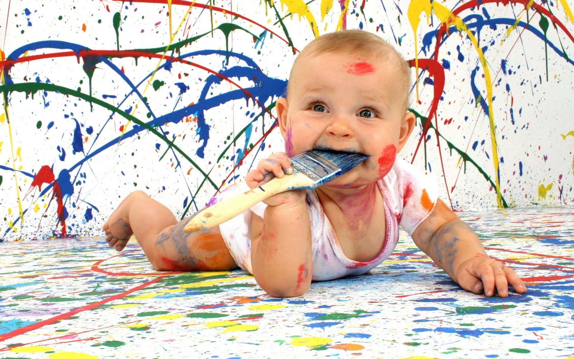 A little kid all in paint, licking with a brush smiling