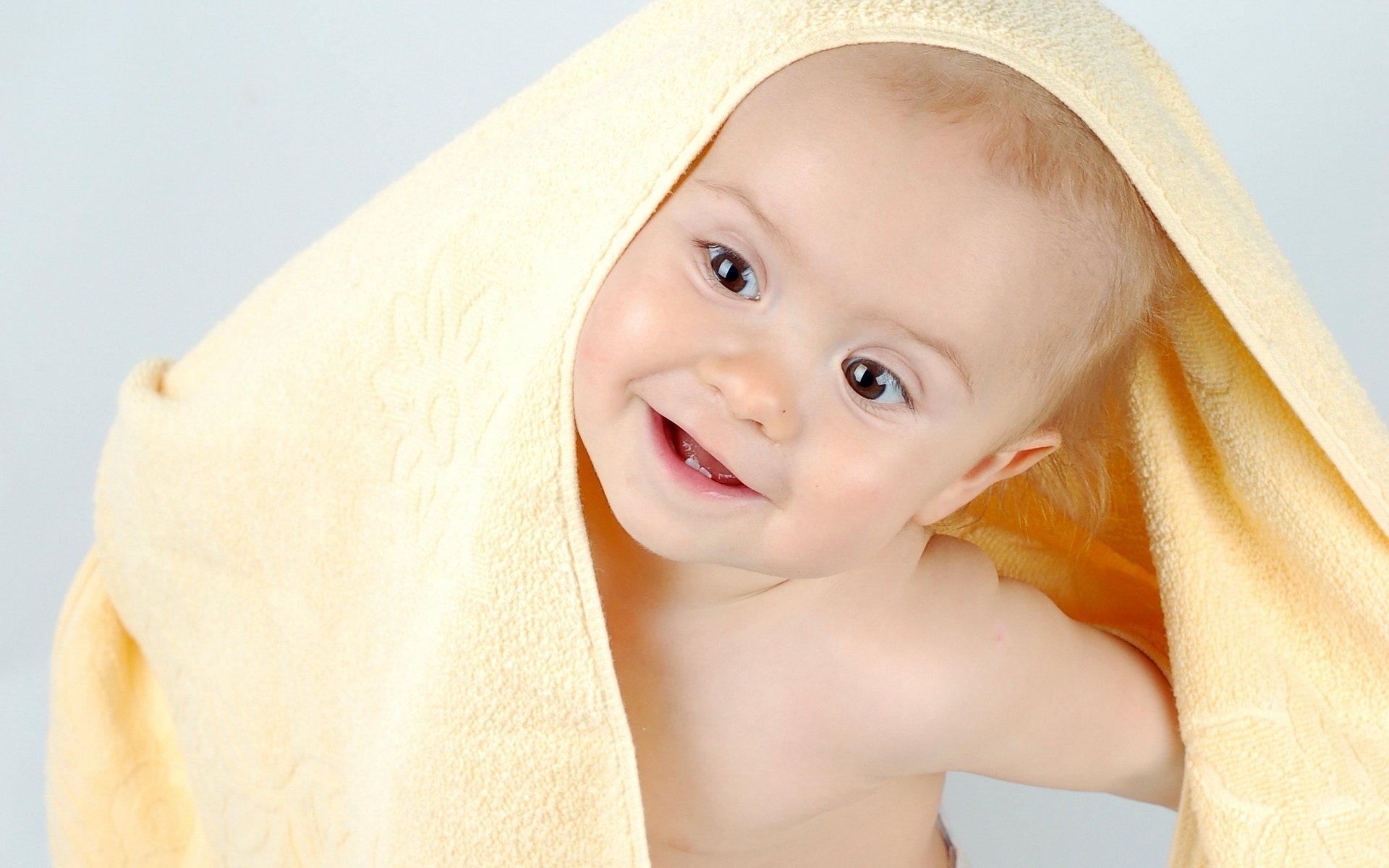 A baby peeking out from under a yellow terry towel